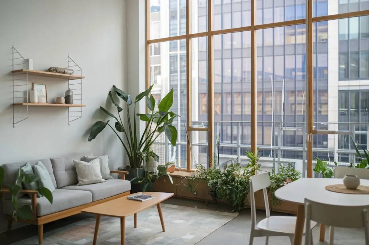Un salon lumineux avec une grande fenêtre, des plantes vertes, un canapé gris et un mobilier moderne en bois clair.