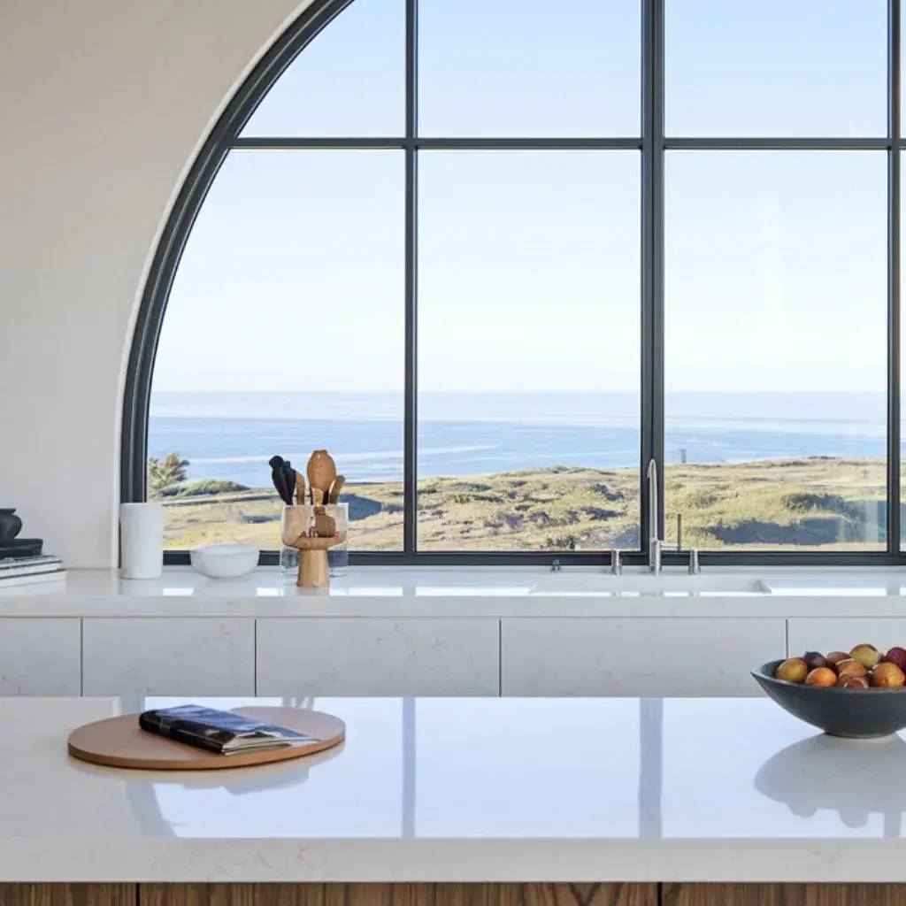 Une élégante cuisine moderne avec vue panoramique sur l'océan, mettant en valeur un grand évier sous une fenêtre voûtée et un plan