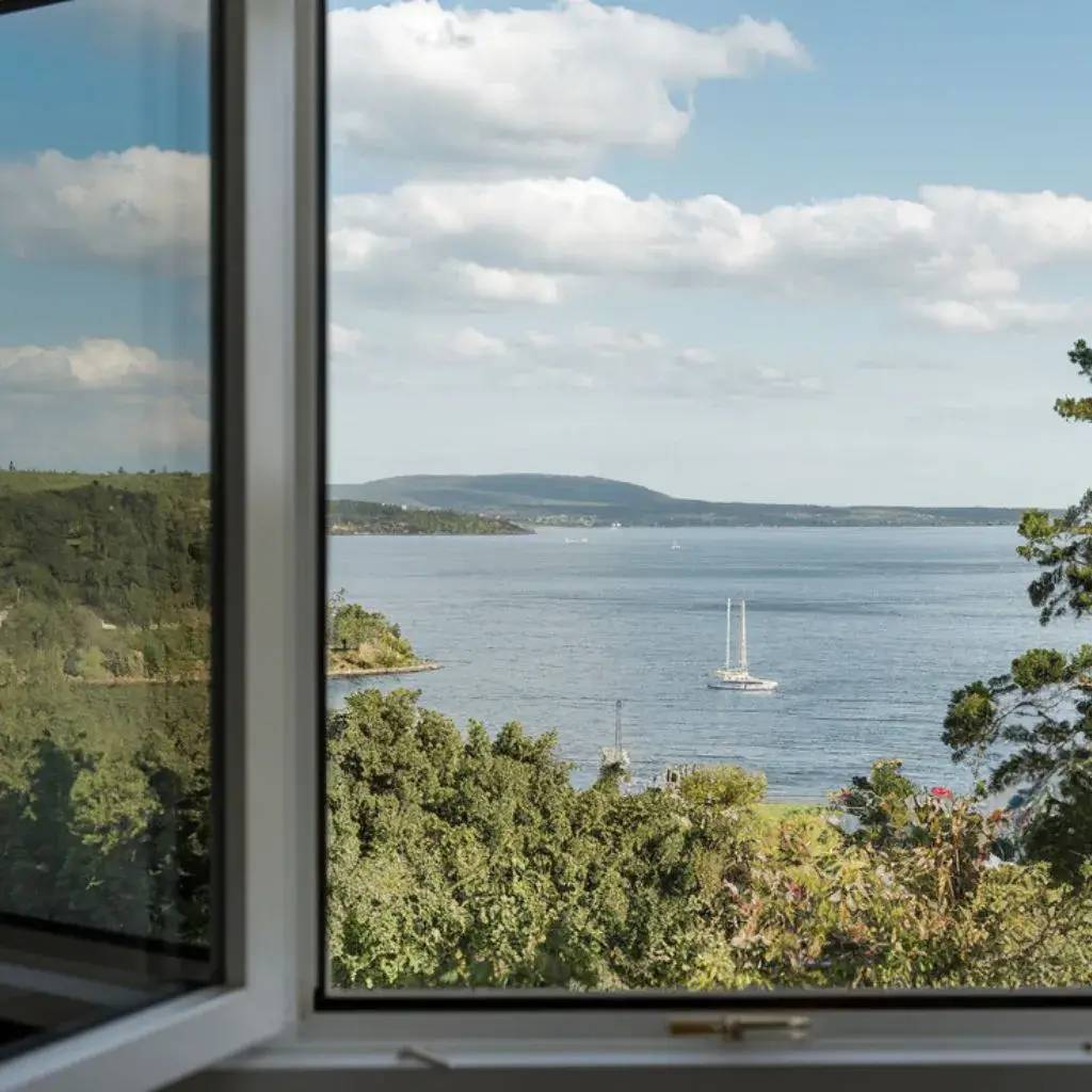 Vue pittoresque d'une fenêtre ouverte donnant sur un paysage côtier avec un voilier et une végétation verdoyante.