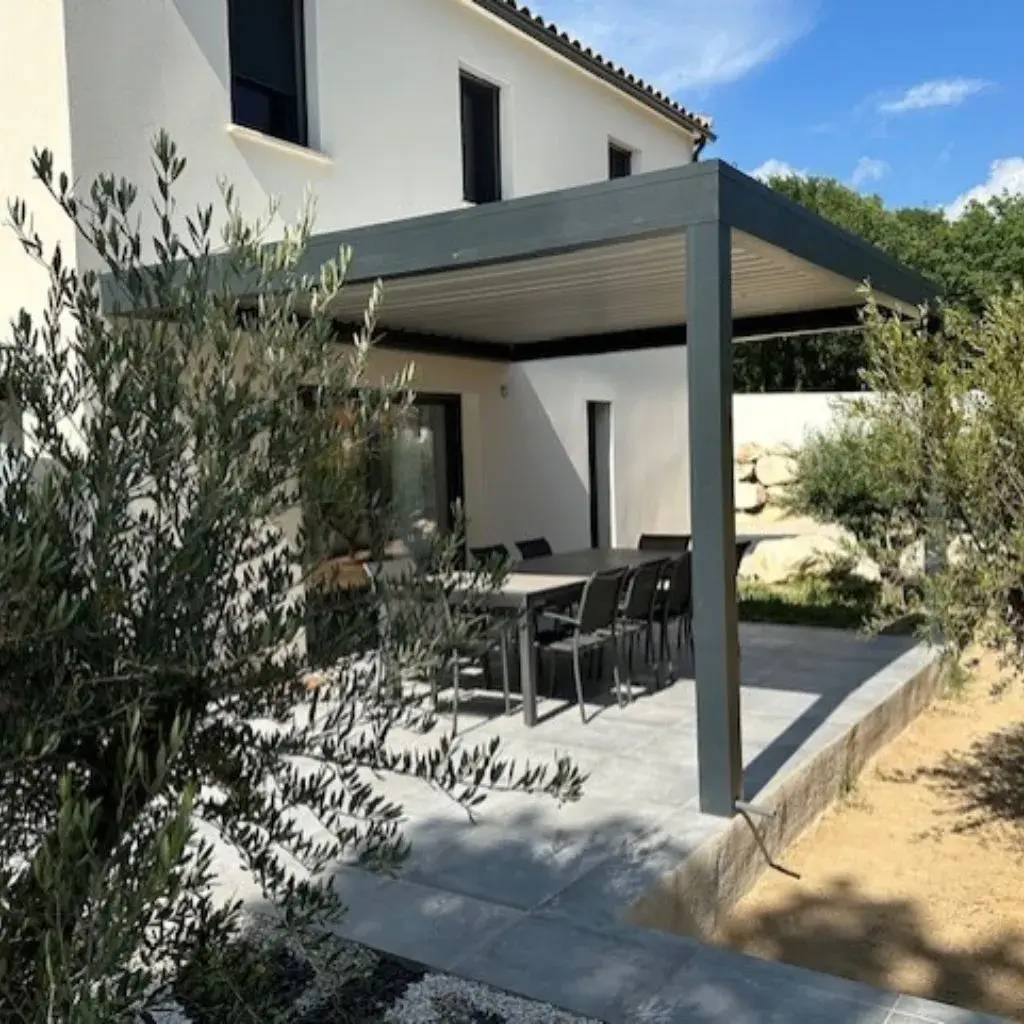 Une terrasse moderne avec un auvent en métal abrite une grande table entourée de chaises, entourée de verdure.