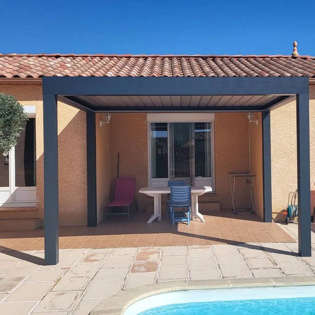 Une terrasse ombragée avec un toit moderne surplombe un coin détente en bord de piscine, devant une maison au toit de tuiles.