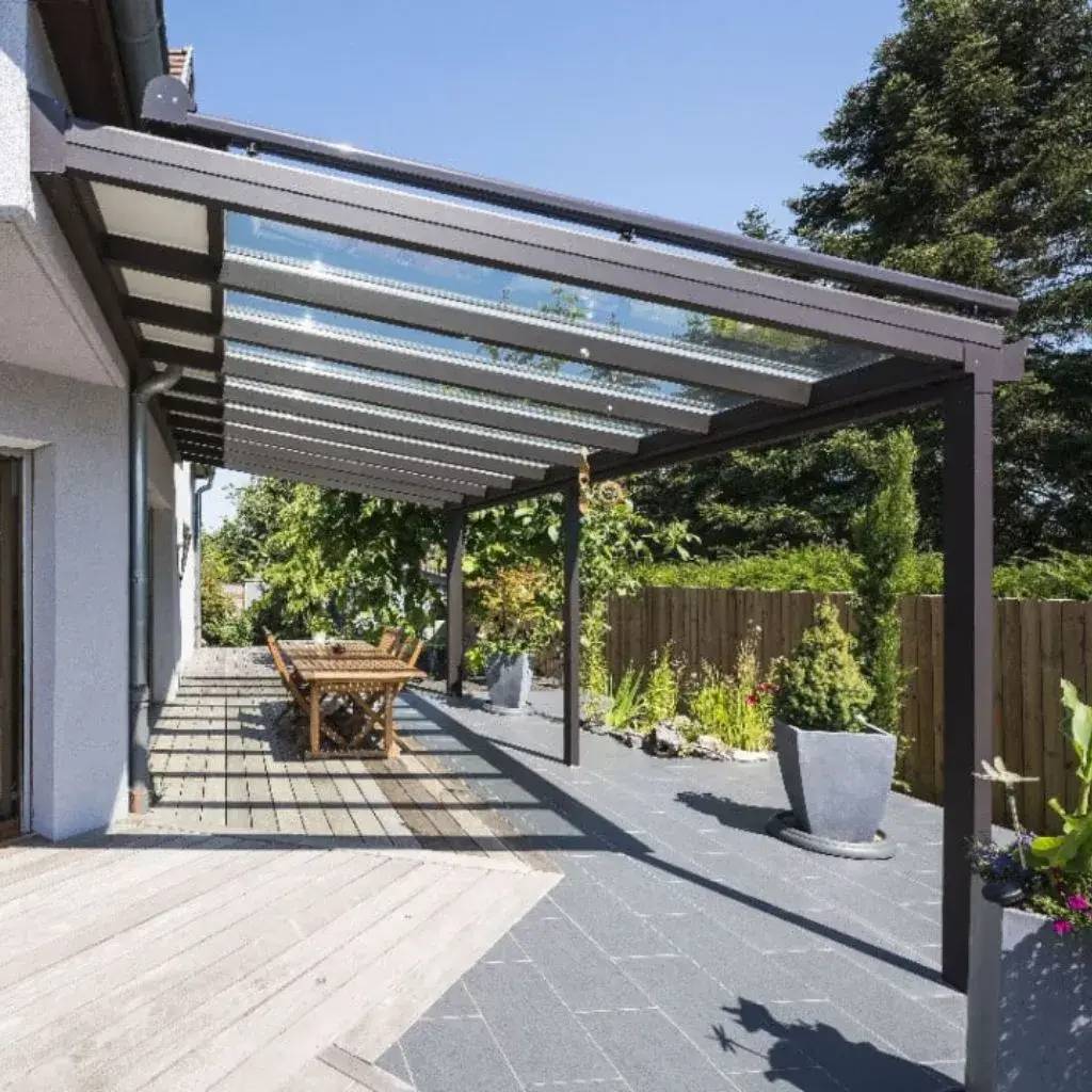 Une terrasse moderne avec un toit en verre, équipée d'un mobilier en bois, bordée de plantes vertes et baignée de lumière naturelle.
