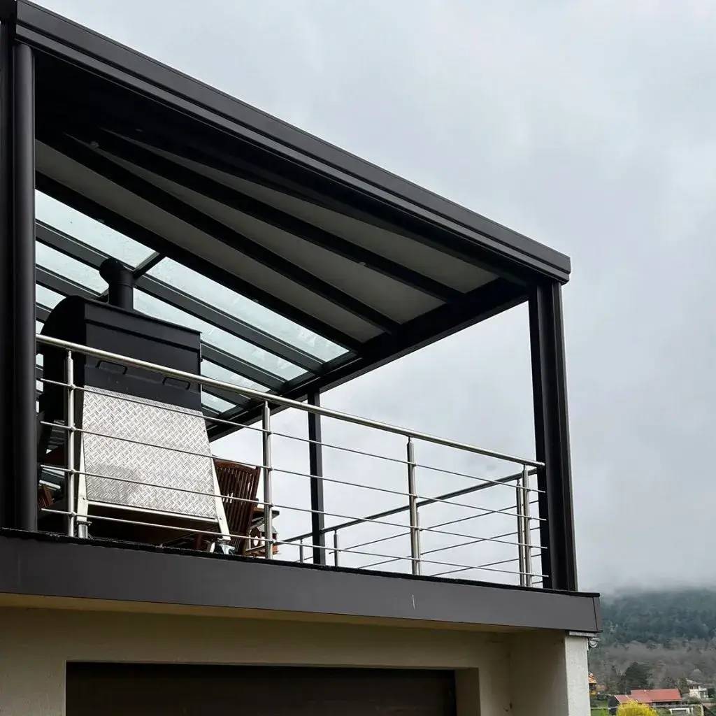 Un balcon moderne avec un toit en verre et garde-corps en métal, abritant un petit meuble et un poêle.