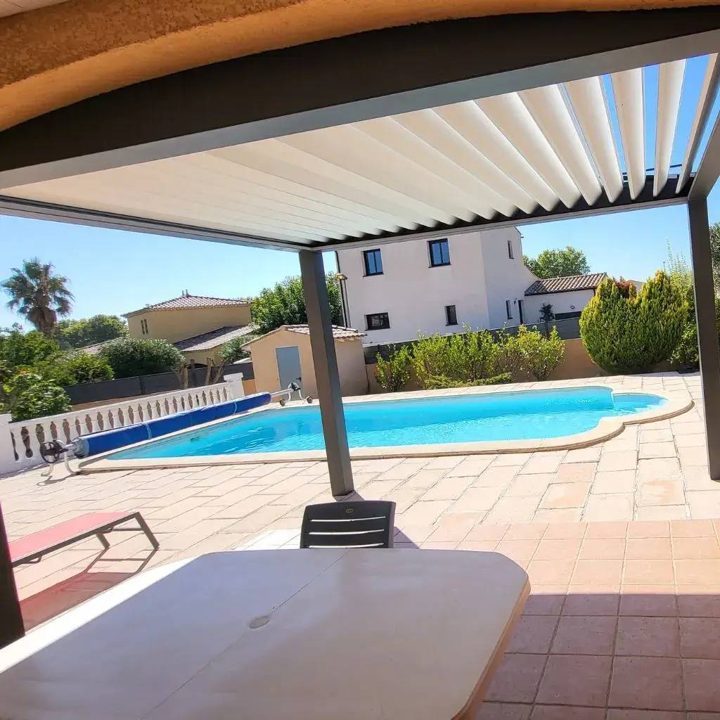 Une terrasse ombragée donnant sur une piscine ensoleillée entourée de maisons, parfaite pour se détendre.