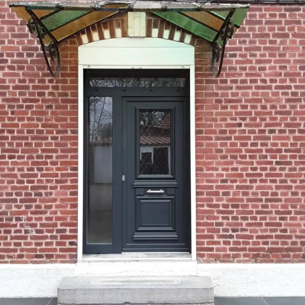 Porte moderne en bois sombre avec verrière, encadrée de briques rouges et abritée par un auvent décoratif en fer forg