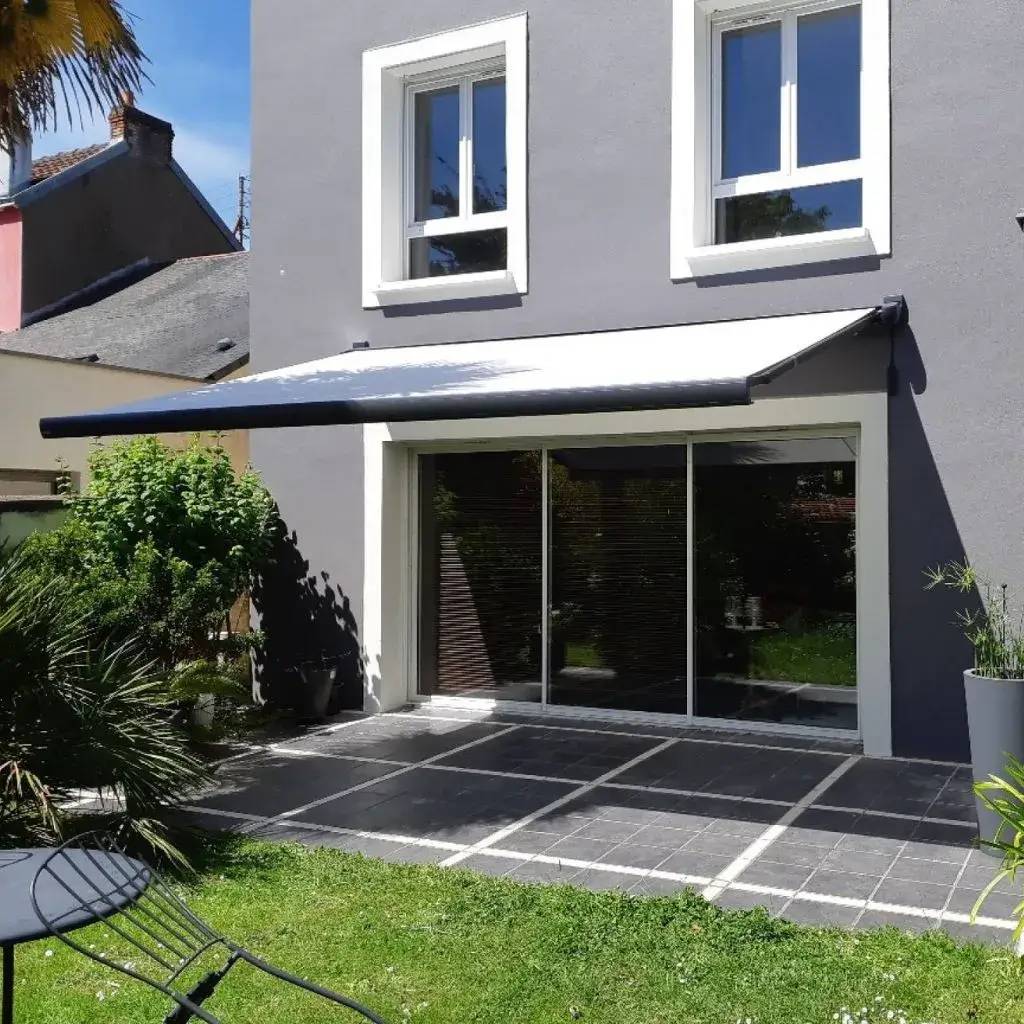 La photo montre une terrasse ensoleillée avec un store banne moderne étendu devant une maison aux murs gris et aux bordures blanches.