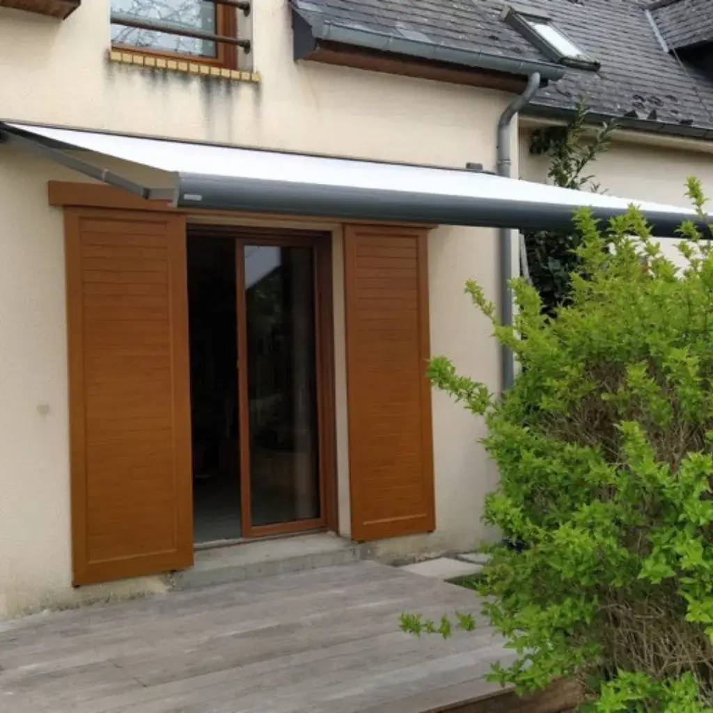 Une terrasse avec une porte-fenêtre en bois et un store rétractable, entourée de verdure.