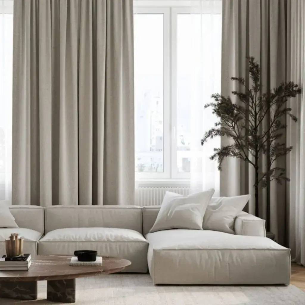 Un salon moderne et épuré avec un grand canapé beige, des rideaux épais et une table basse en bois, baigné de lumière naturelle.