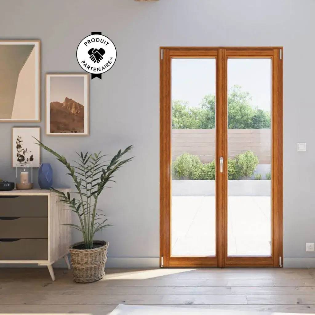 Une porte-fenêtre en bois donne sur une terrasse ensoleillée, entourée de verdure, avec une plante d'intérieur et des photos enc