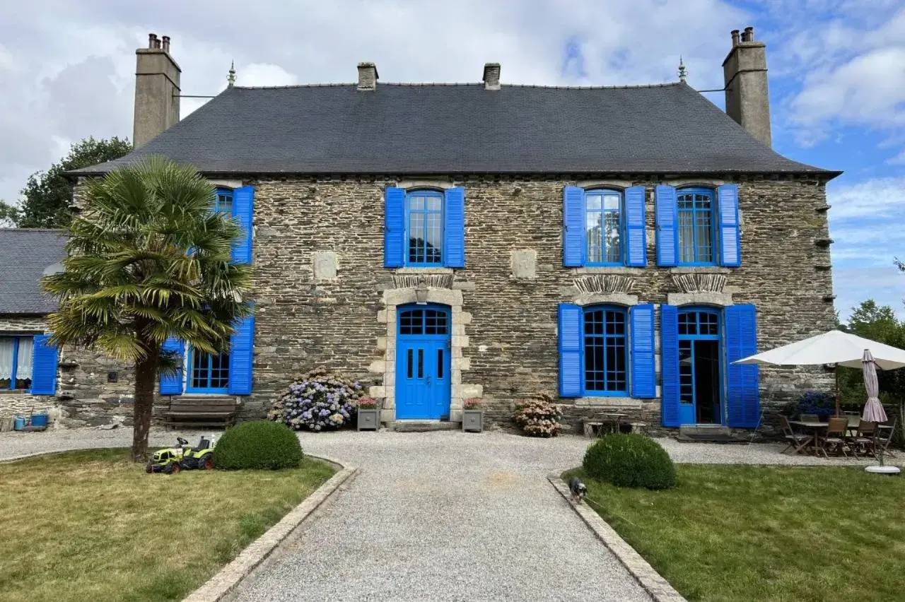 Maison en pierre avec volets et porte bleus, entourée de verdure et d'un jardin aménagé, sous un ciel partiellement nuage