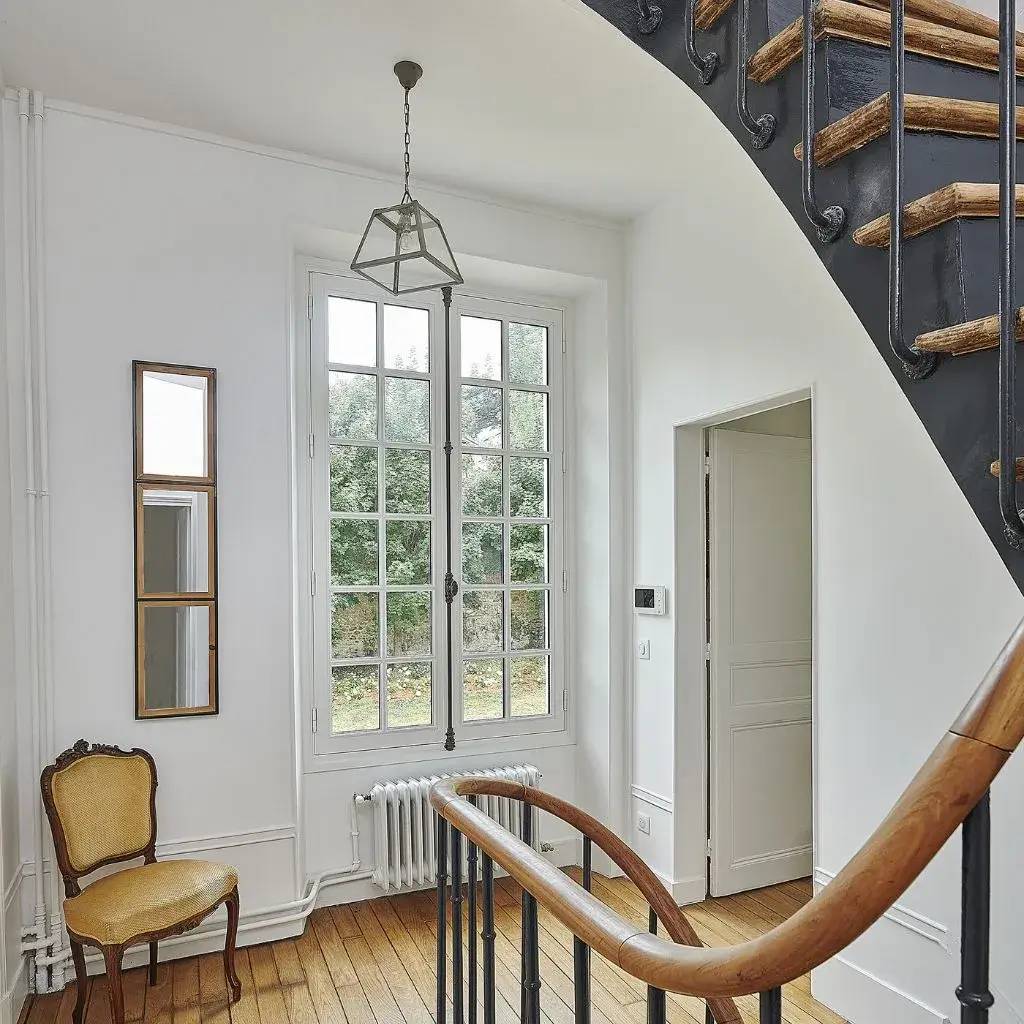 Un hall d'entrée lumineux avec un escalier élégant, une fenêtre à grandes vitres, un radiateur moderne et une chaise classique.