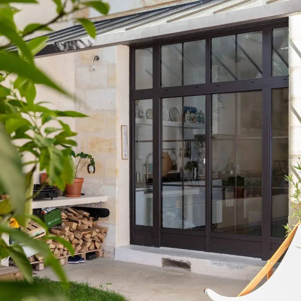 Une grande baie vitrée en métal noir s'ouvre sur une cuisine moderne, entourée de murs en pierre claire, avec des plantes et une pile