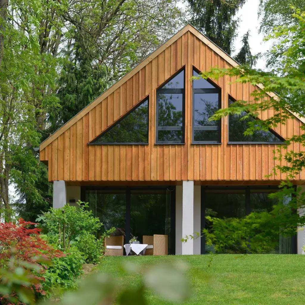 Maison moderne en bois avec de grandes fenêtres triangulaires, entourée de verdure luxuriante.