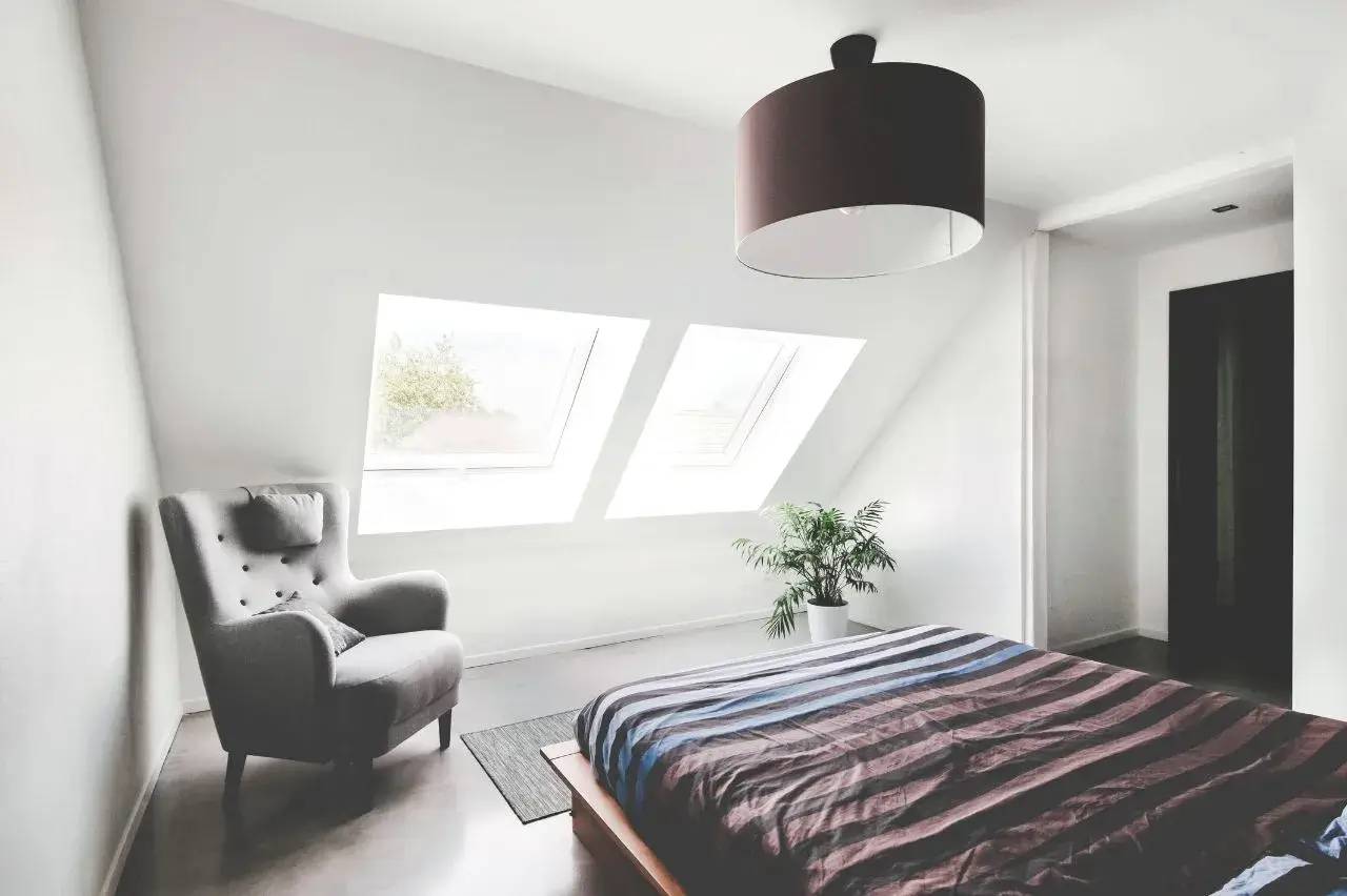 Une chambre lumineuse avec un fauteuil gris, un lit à rayures bleues et bordeaux, deux grandes fenêtres de toit, et une plante verte
