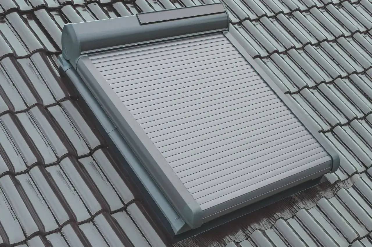 Un volet roulant métallique installé sur une fenêtre de toit en tuiles grises.