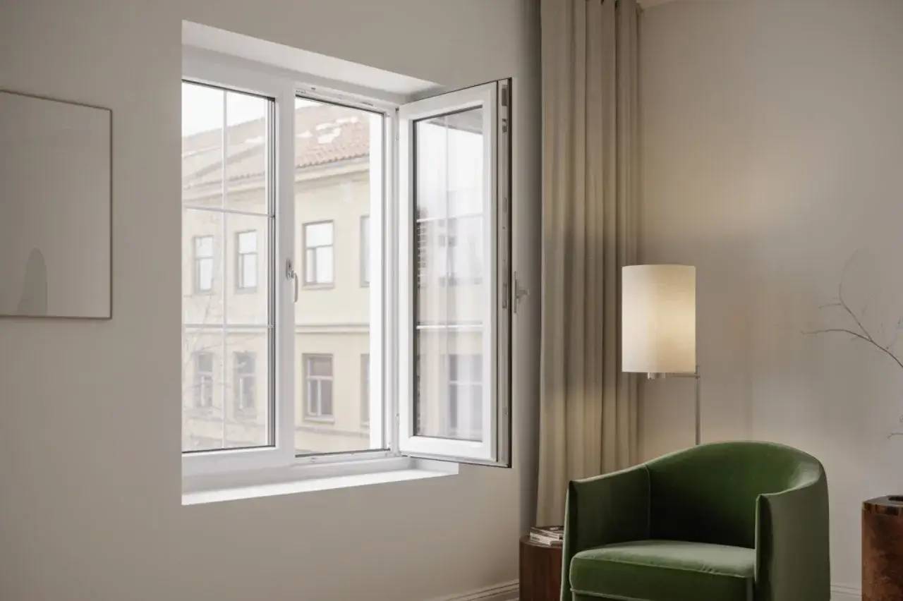 Une fenêtre ouverte laisse entrer la lumière naturelle dans un intérieur moderne et épuré, agrémenté d'un fauteuil vert et d'une lampe sur pied.