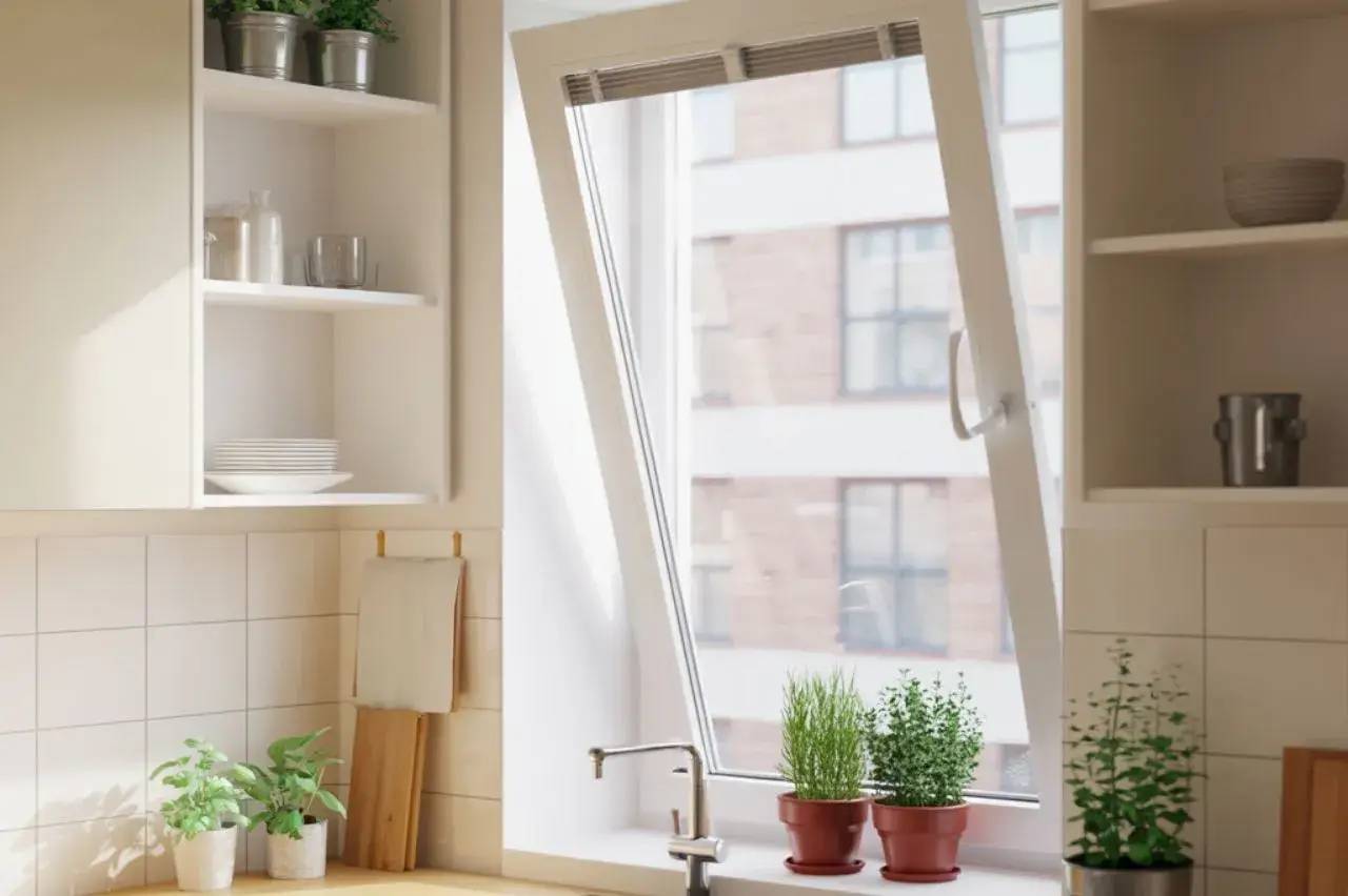 Une cuisine lumineuse avec des plantes en pot sur le rebord d'une fenêtre ouverte et des étagères blanches garnies de vaisselle.