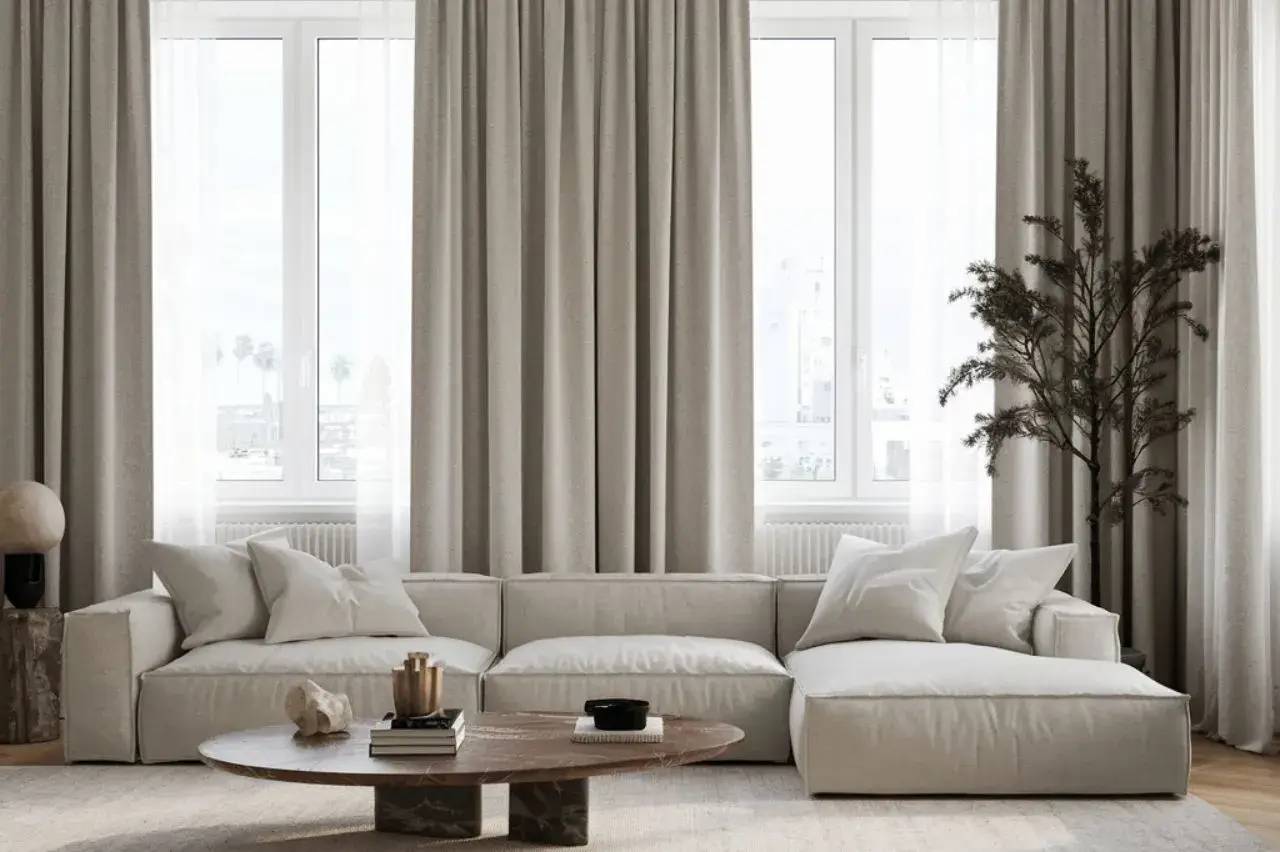 Un salon lumineux et moderne avec un canapé beige, une table basse en bois et des rideaux légers devant de grandes fenêtres.