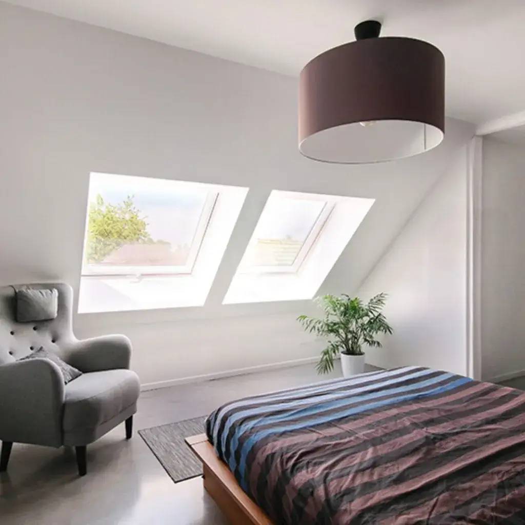 Une chambre moderne et lumineuse sous combles, avec de grandes fenêtres inclinées, un fauteuil gris, une plante en pot et un lit à ray