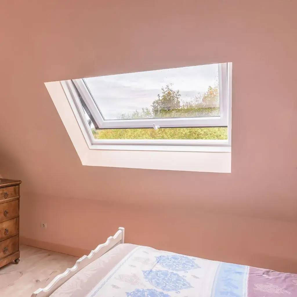 Une chambre lumineuse aux tons doux, avec une fenêtre de toit ouverte offrant une vue sur des arbres.