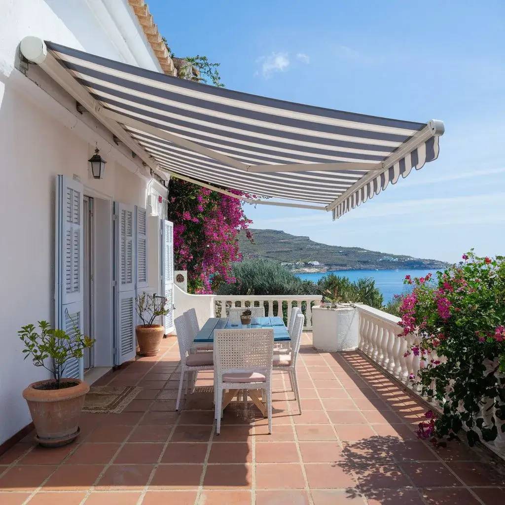 Une terrasse ensoleillée avec une vue sur la mer, ornée de fleurs colorées et d'un store rayé gris et blanc.