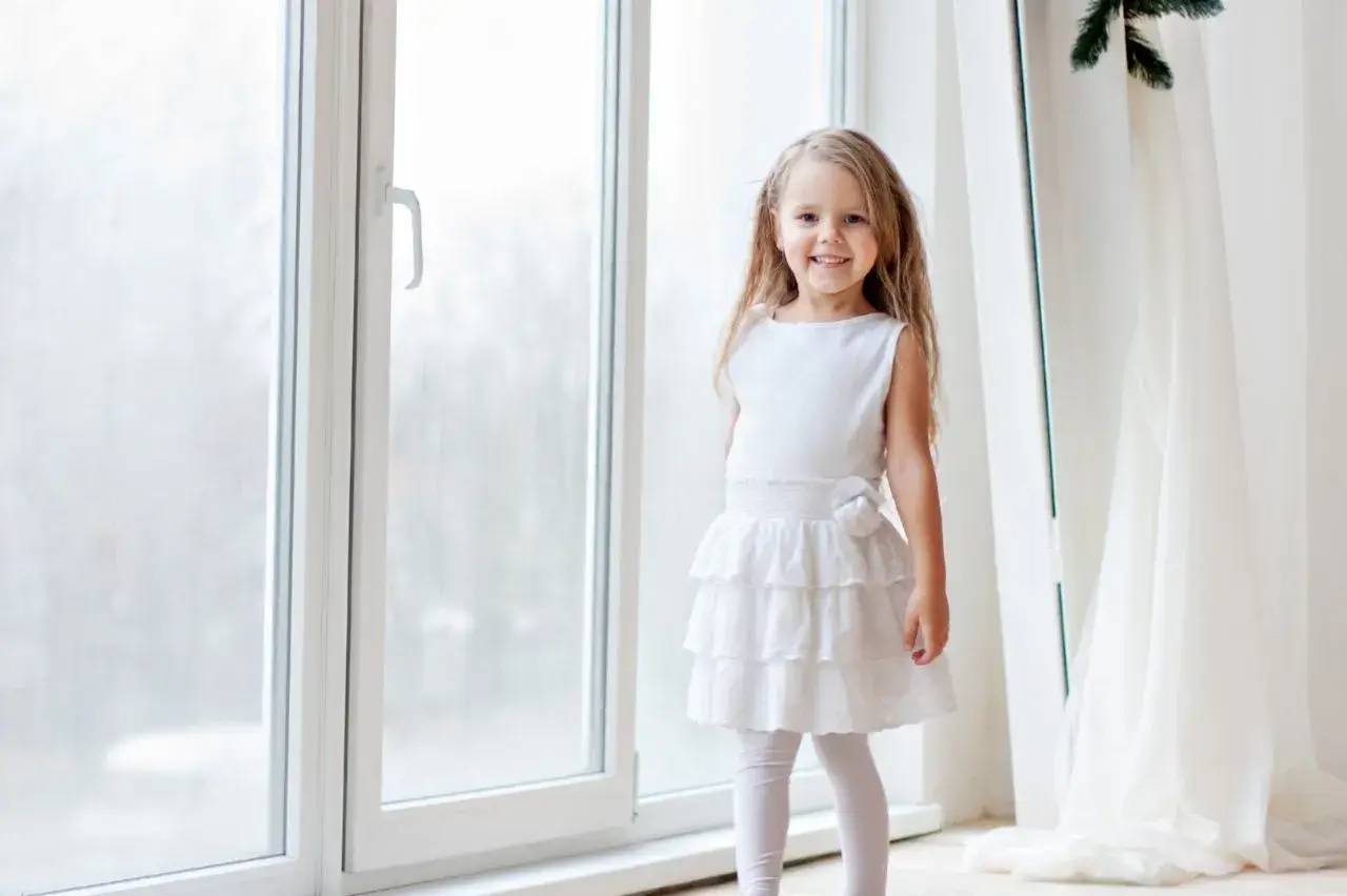 Une petite fille souriante en robe blanche se tient debout près d'une grande fenêtre.