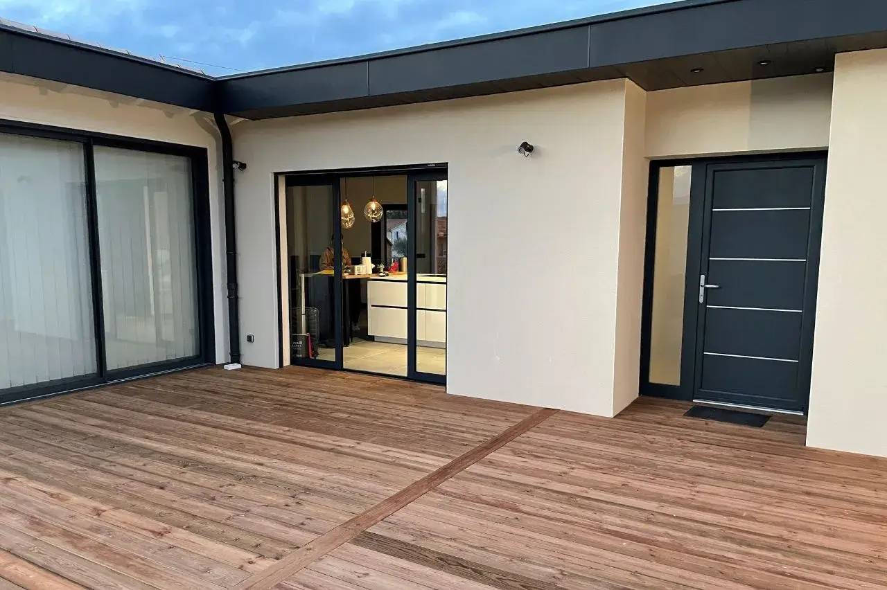 Une terrasse en bois moderne jouxte une maison aux grandes baies vitrées et porte d'entrée élégante.