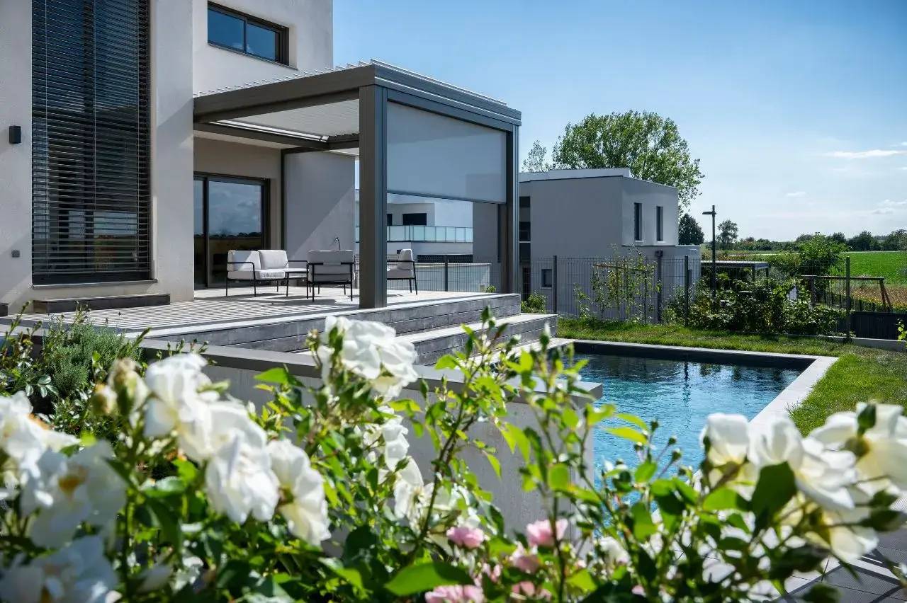 Cette image montre une terrasse moderne surplombant un jardin fleuri avec piscine, sous un ciel ensoleillé.