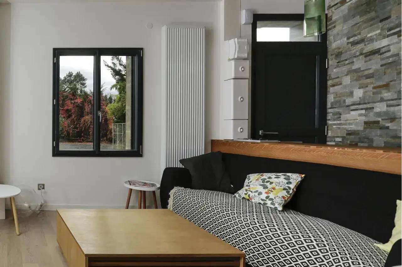 Un salon moderne et épuré avec un canapé noir, des coussins colorés, une table basse en bois clair, et une fenêtre offrant une vue