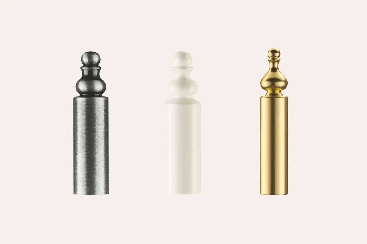 Trois bouchons cylindriques en métal de couleurs différentes (gris, blanc et doré) sont alignés sur un fond beige.