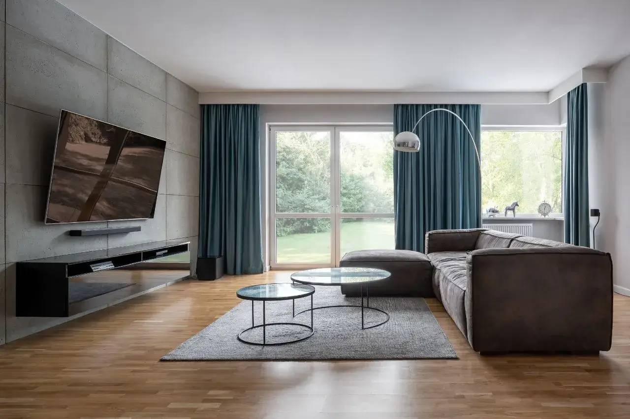 Un salon moderne et épuré avec un canapé d'angle gris, deux tables basses en verre sur un tapis, et une grande télévision murale