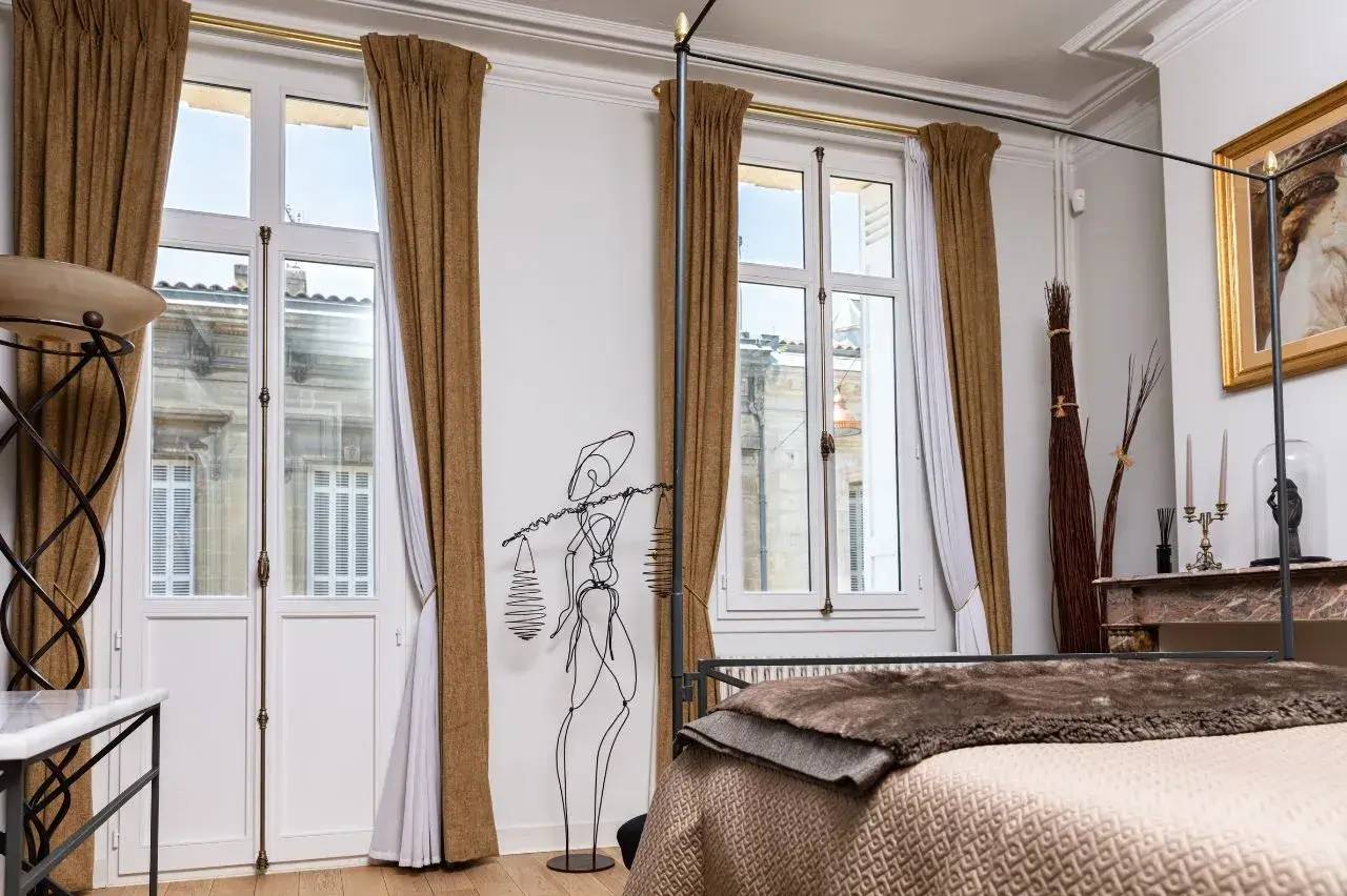 Une chambre élégante avec de grandes fenêtres encadrées de rideaux marron, une sculpture métallique stylisée et une cheminée décorée.