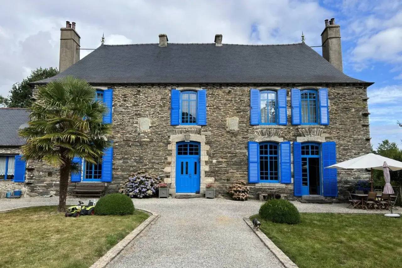 Une maison en pierre avec des volets bleus vifs et un jardin bien entretenu, sous un ciel partiellement nuageux.