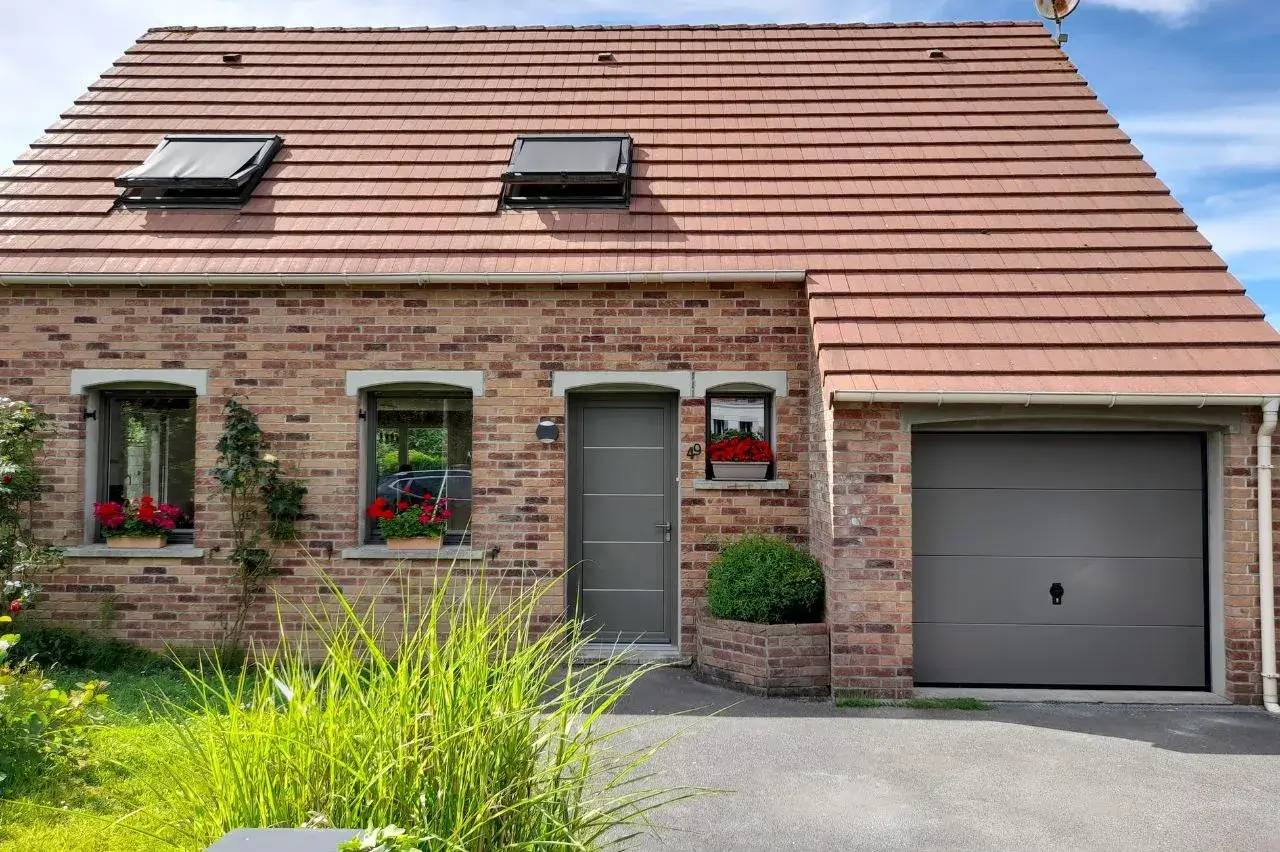 Façade de maison en briques rouges avec toit en tuiles, fenêtres ornées de fleurs rouges, et porte de garage grise.