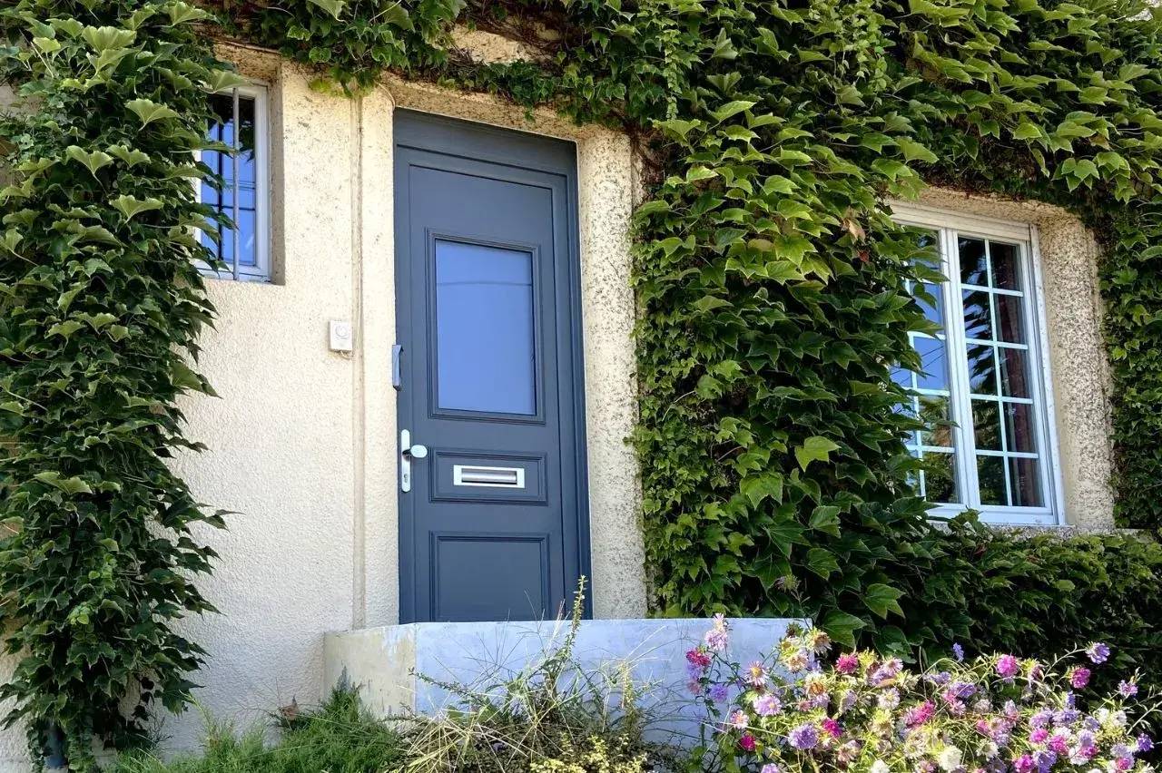 Une porte d'entrée bleue entourée de murs recouverts de lierre, avec une petite fenêtre à gauche et des fleurs colorées au premier