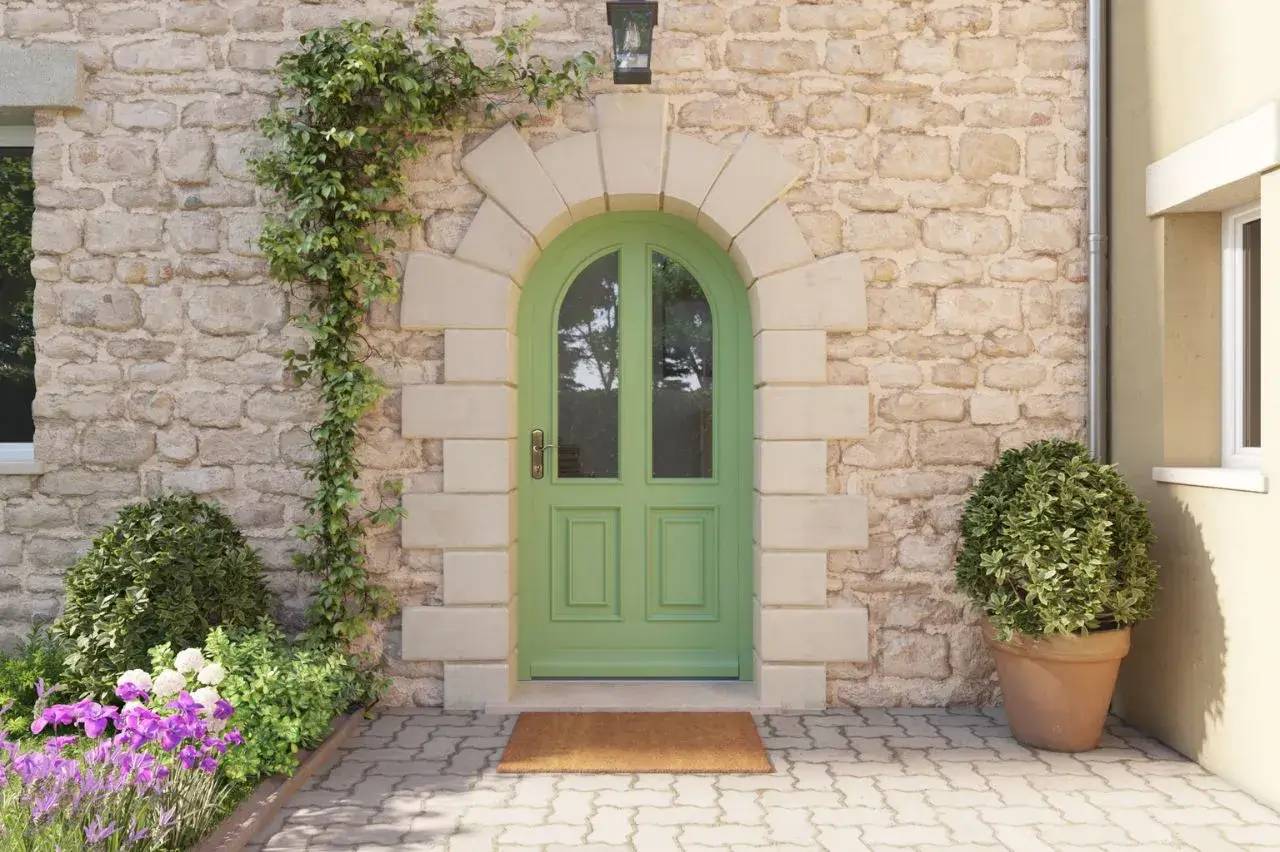 Une porte verte arrondie, encadrée de pierre claire sur un mur en pierre, est flanquée de plantes grimpantes et de