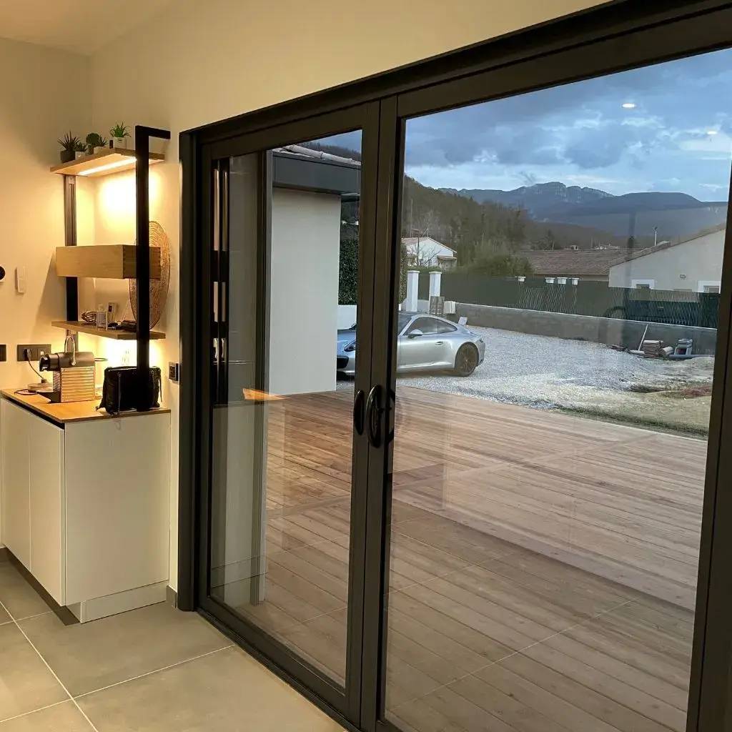 La photo montre une cuisine moderne avec une grande baie vitrée donnant sur une terrasse et un paysage montagneux.