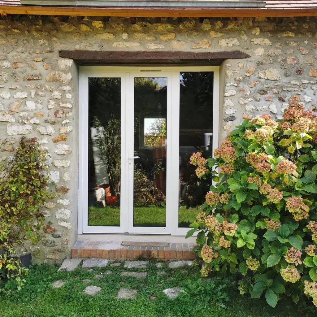 Une porte vitrée moderne est encadrée par un mur en pierre avec des plantes et des fleurs à proximité.