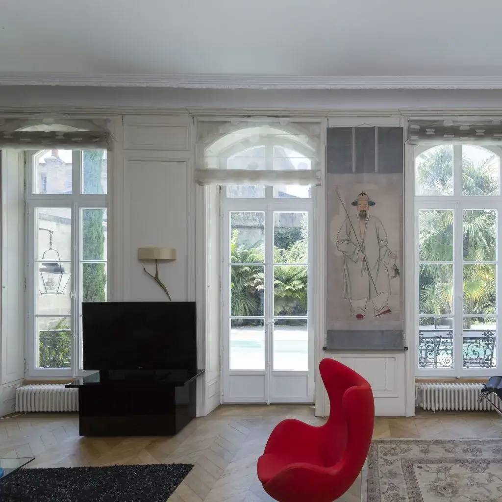 Une pièce lumineuse et élégante avec de grandes fenêtres donnant sur un jardin, une chaise rouge moderne et une œuvre d'art orientale accrochée