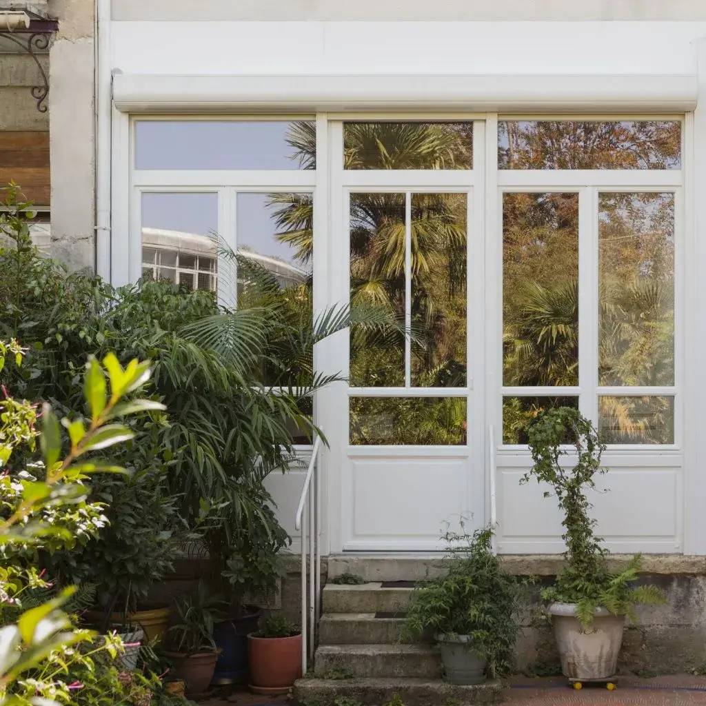 Une façade lumineuse avec de grandes fenêtres blanches entourées de plantes vertes luxuriantes.