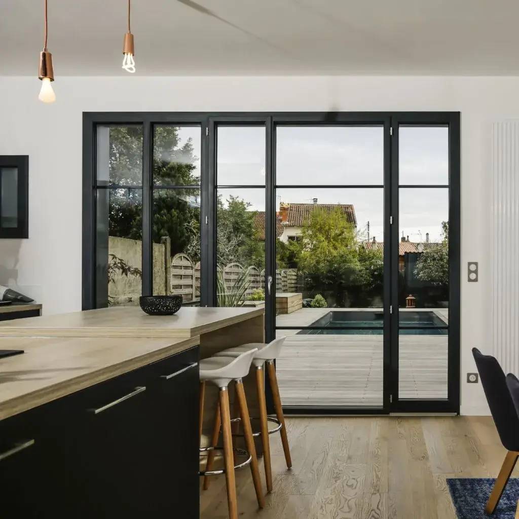 Une cuisine moderne avec un îlot central en bois et des tabourets, donnant sur une terrasse avec piscine à travers de grandes baies vitrées