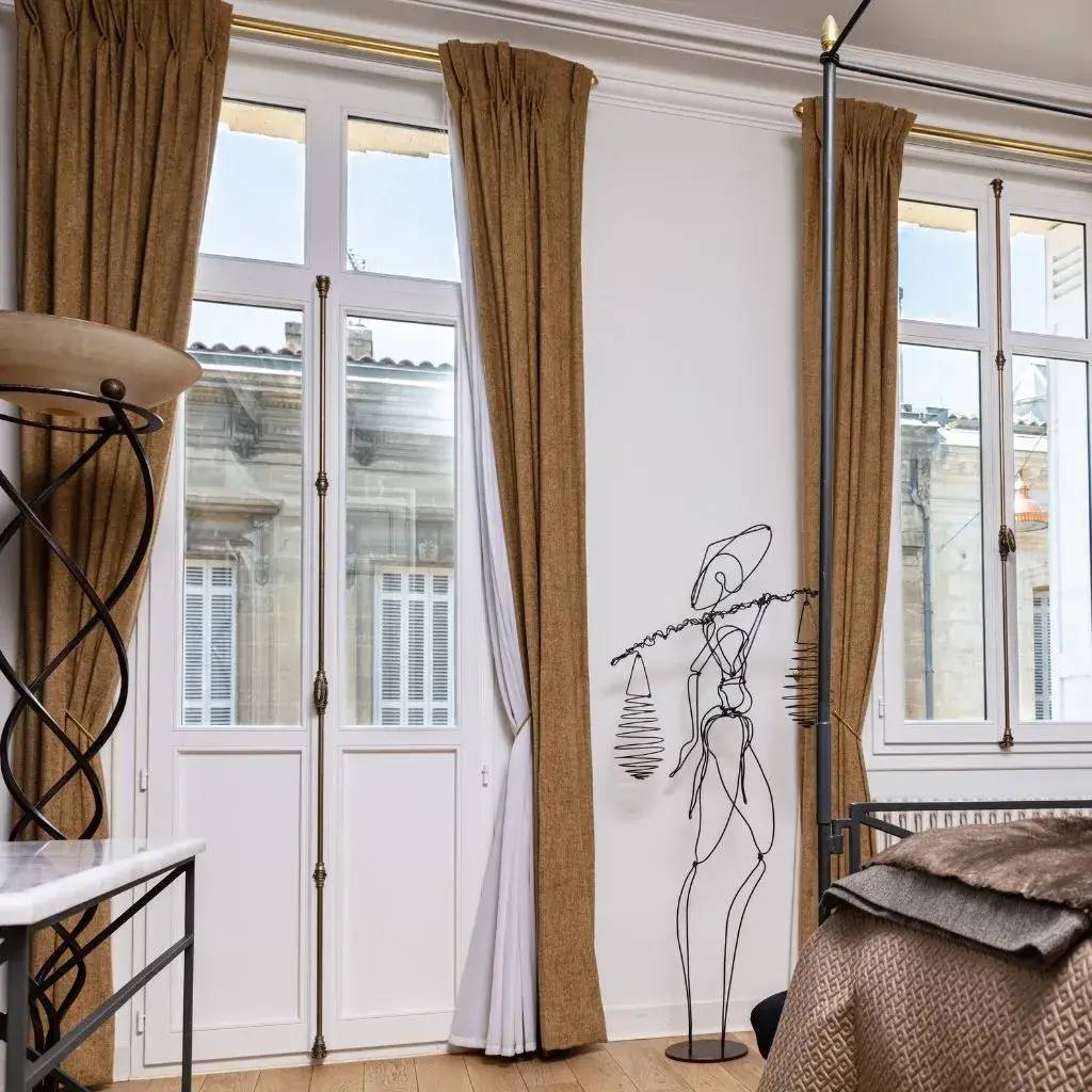 Cette image montre une chambre élégante avec de grandes fenêtres, des rideaux beiges et une sculpture métallique minimaliste.