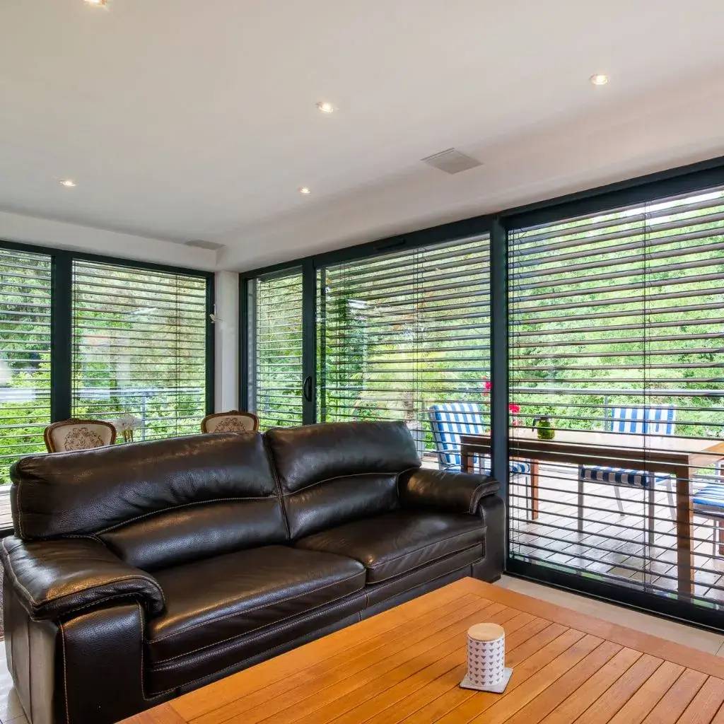 Un salon moderne avec un canapé en cuir noir, doté de grandes baies vitrées et offrant une vue sur une terrasse avec des meubles extérieurs.