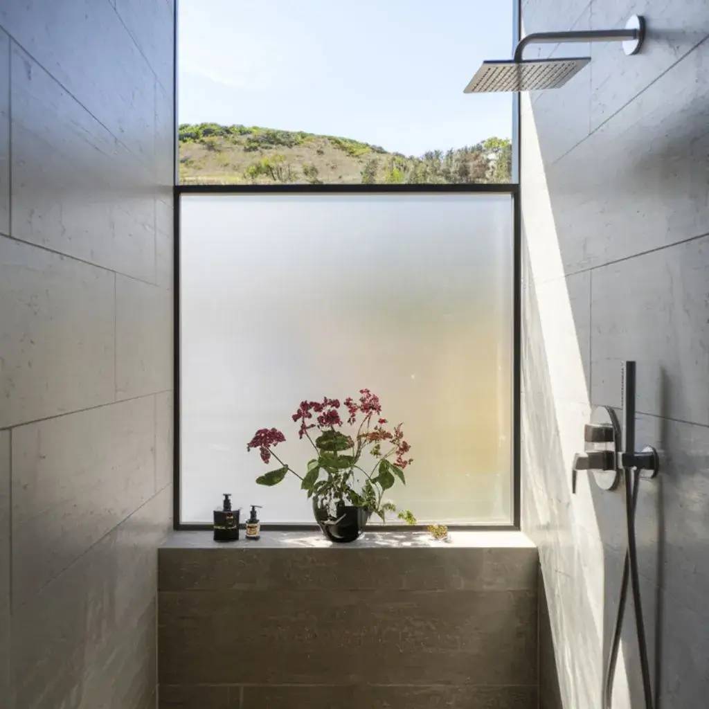 Douche moderne avec une grande fenêtre dépolie encadrant un vase de fleurs rouges sur une niche en pierre.