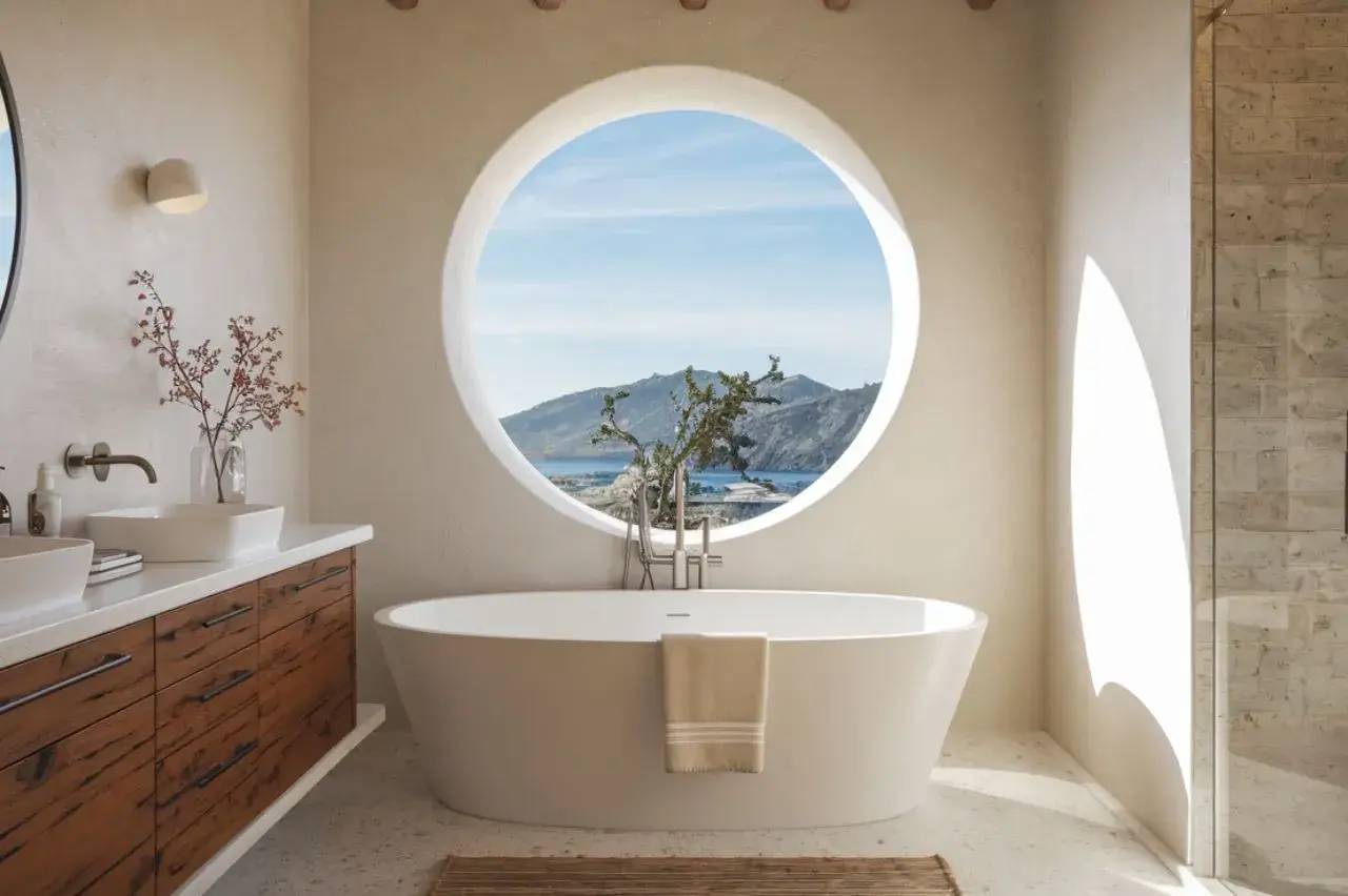 Une salle de bain lumineuse avec une baignoire ovale et une grande fenêtre ronde offrant une vue panoramique sur la mer.