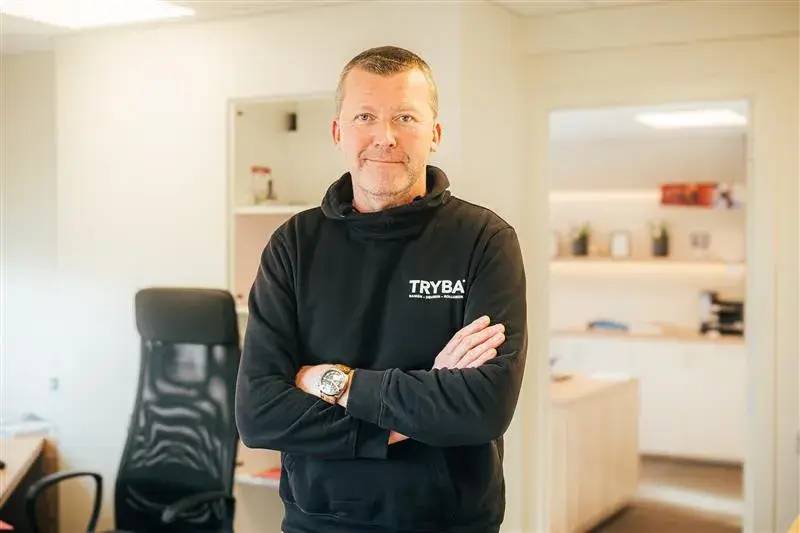 Un homme en sweat-shirt "TRYBA" se tient avec les bras croisés dans un bureau lumineux et moderne.
