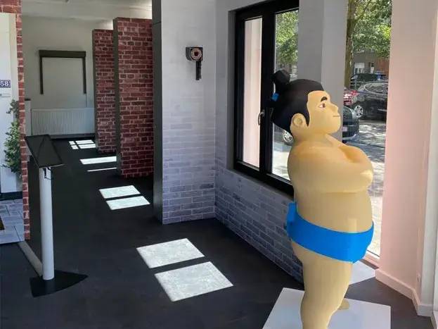 Une statuette colorée de sumo en tenue traditionnelle bleue se tient près d'une fenêtre dans une pièce aux murs de briques et carrel