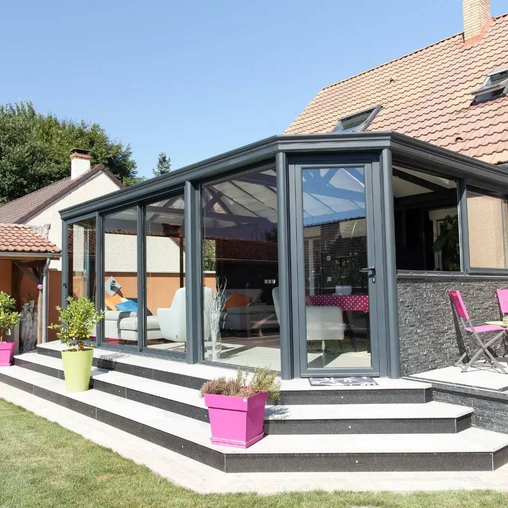 Une véranda moderne en verre et métal gris, agrémentée de pots de fleurs colorés, s'ouvre sur un jardin verdoyant.