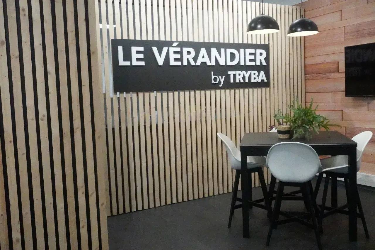 Espace moderne avec un panneau "LE VÉRANDIER by TRYBA", des chaises blanches autour d'une table noire, et une décoration