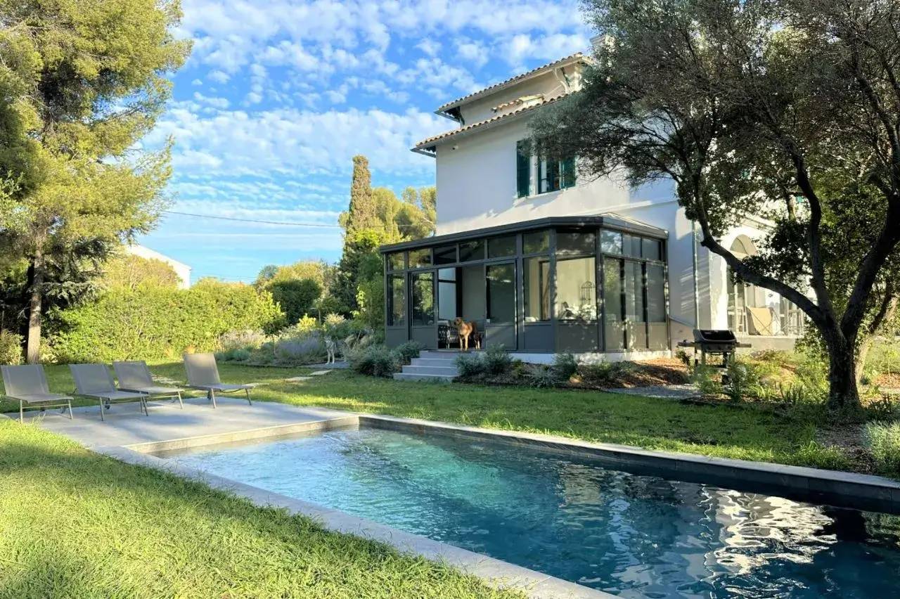 Grande maison blanche avec véranda, entourée d'un jardin verdoyant et d'une piscine moderne.